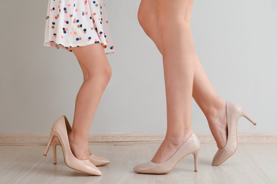 Legs of young mother with her cute little daughter wearing high-heels near light wall