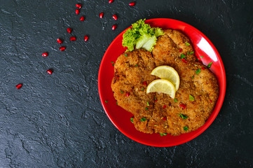 Large Viennese schnitzel on a red plate with lemon on a black background. Meat dish. Top view, flat...