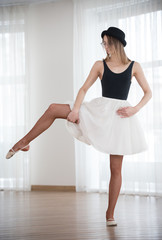 Ballerina in a hat bends a leg to the side, in the studio