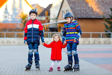 Two kids boys and little toddler girl playing together outdoors on sunny day. Brothers in protection safety clothes skating with rollers. Happy sister holding siblings. Happy family of three children