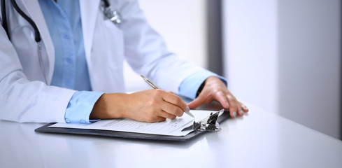 Unknown doctor woman filling up medical form while sitting at the desk in hospital office. Physician at work. Medicine and health care concept