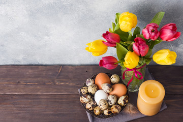Concept Easter, tulips in a vase and a basket of eggs, a candle on a gray background. Copy space for product or text.