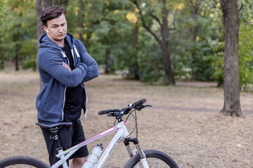 Young handsome caucasian man stands hands crossed with  bicycle frowning and serious. White wireless earphones, casual sportswear. Abandoned park background. Outdoor, copy space.