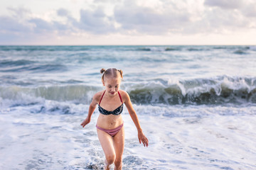 Fototapeta premium Blonde girl running and jumping on the beach on blue sea shore in summer vacation at the day time. Blue ocean with white big wawes on the background