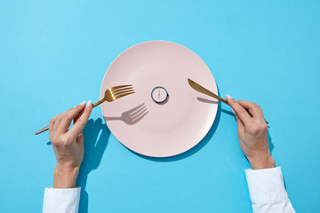 White plate with round whatch shows six o'clock served knife and fork in a girl's hands on a blue...