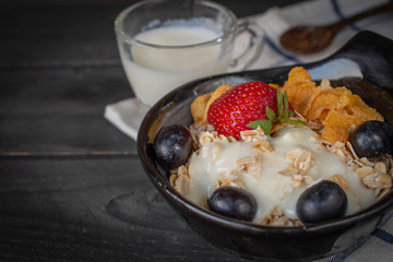 Yoghurt mix oatmeal, strawberry and grape topping in black bowl on Blue and white striped fabric and wood table with spoon and milk in glass placed back side.