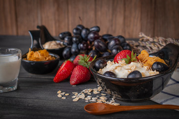 Yoghurt mix oatmeal, strawberry and grape topping in black bowl on wood table with Blue and white striped fabric, spoon, strawberry, milk in glass, grape, cornflakes and oatmeal placed around.