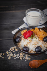 Yoghurt mix oatmeal, strawberry and grape topping in black bowl on wood table with Blue and white striped fabric, spoon, oatmeal and milk in glass placed around.
