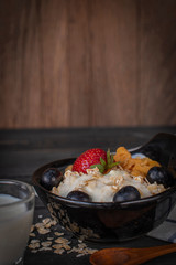 Yoghurt mix oatmeal, strawberry and grape topping in black bowl on wood table with Blue and white striped fabric, spoon, oatmeal and milk in glass placed around.