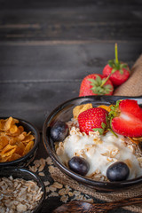 Yoghurt mix oatmeal, strawberry and grape topping in black bowl on sack and wood table with spoon, oatmeal, cornflakes and strawberry placed around.