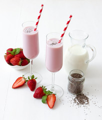 Strawberry smoothie. 2 glasses of strawberry milkshake, glass pitcher of milk, small jar of chia seeds, porcelain bowl with strawberries embellished with few real strawberries and fresh mint.