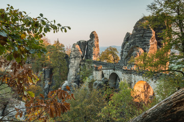 Bastei bridge in the evening sun in the national park Saxon Switzerland. Elbe sandstone mountains with trees in autumn colors and rock formations and old tree trunk and leaves in the foreground