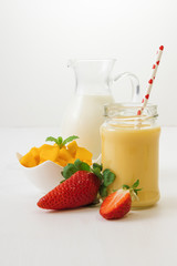 Glass of fresh mango smoothie or mango milkshake with milk in glass pitcher, pieces of mango in porcelain bowl and few real fresh strawberries on white wooden table. Closeup view.