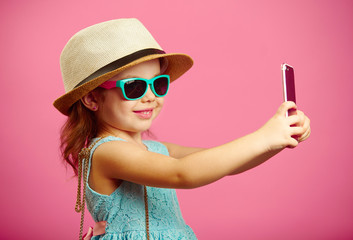 Beautiful little girl dressed in summer dress, wearing a straw hat and sunglasses, makes a selfie...