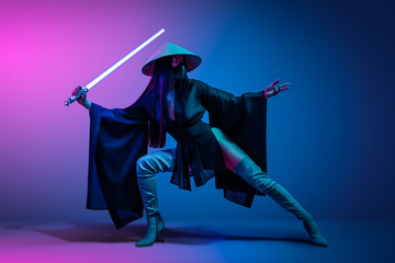 Concept on cosmic cosplay. Сontemporary portrait a young athletic woman in traditional Japanese black kimono, an Asian hat and highboots is holding a lightsaber and posing on neon blue-pink background - obrazy, fototapety, plakaty