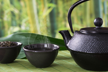 Asian green tea set with black china kettle on bamboo mat with dried green tea in bowl. Chinese tea...