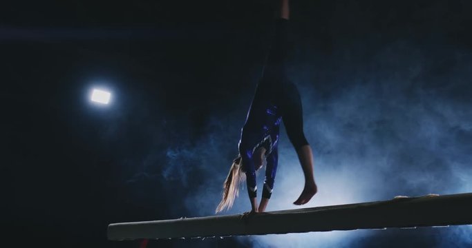 The girl is a professional athlete performs gymnastic acrobatic trick on a beam in backlight and slow motion in sports gymnastic clothing. Smoke and blue. Jump and spin on the balance beam