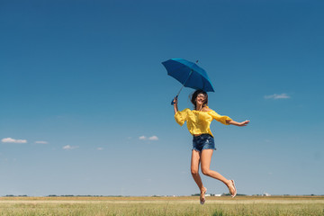 Beautiful Caucasian girl in yellow blouse and denim shorts jumping with blue umbrella outdoors....