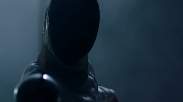 Professional fencer in protective uniform practicing maneuvers with sword alone in dark studio . Fencing demonstration on black smoked background . Shot on Arri Alexa cinema camera in slow motion .