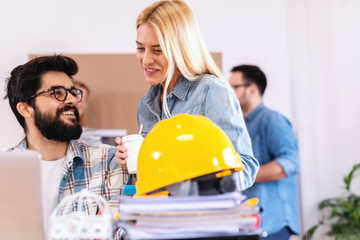 Two architects looking at laptop and developing project. Blonde woman smiling ad holding coffee to go while bearded man sitting and looking at her. Start up business concept.