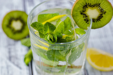 Summer refreshing drink in a glass with straw close-up. Cold sweet and sour lemonade with cubes of lemon, kiwi, mint and ice on a gray wooden table.