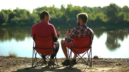 Childhood friends sitting on river bank, drinking beer and discussing fishing