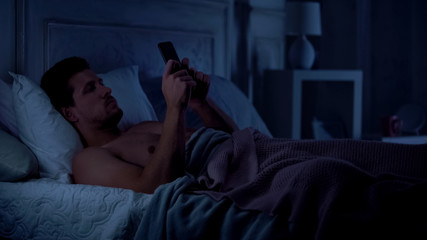 Man lying in bed and surfing net, scrolling social networks, gadget addiction