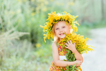 portrait of a little blond blue-eyed smiling girl 4 years on the street in summer, in a rustic style with a wreath of yellow flowers on her head and in a yellow dress