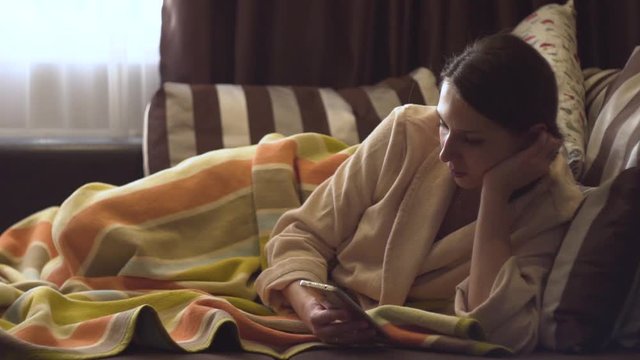 Lady in beige bathrobe with smartphone in the bed.