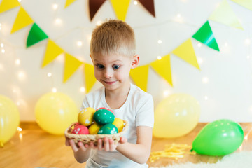 Fototapeta na wymiar blond, blue-eyed cute smiling boy in a white shirt, the age of 6 years, with easter eggs in a room on the background of yellow design, sitting, studio