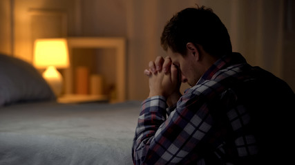 Religious young man praying in evening near bed, belief in God, Christianity - obrazy, fototapety, plakaty