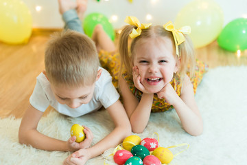 Fototapeta na wymiar blond, blue-eyed cute smiling children,boy in a white t-shirt and ponytail hair girl in yellow dress, brother and sister 4-5-6 years with Easter eggs in a room on the background of yellow decoration