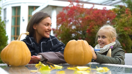 Fototapeta premium Cheerful girl and mom painting scary face on pumpkin, spend time together, fun