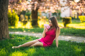 Naklejka na ściany i meble Young girl sitting on the grass and hold a sakura flowers in her hands. Background of pink bokeh of sakura trees
