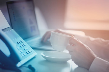 Male hands, cup of coffee, IP phone and laptop on the office table. Ip telephony and video meetings for business concept. Close up.