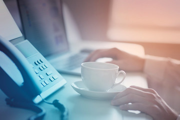 Male hands, cup of coffee, IP phone and laptop on the office table. Ip telephony and video meetings for business concept. Close up.
