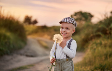 Happy child boy on nature spring,field,meadow,sunlight. Beautiful smiling baby holding big dandelion flower in his hands. Summer portrait little kid wearing fashionable retro clothes, cap,suspenders.