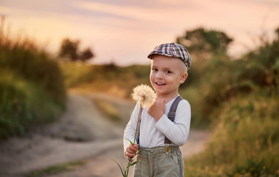 Happy child boy on nature spring,field,meadow,sunlight. Beautiful smiling baby holding big dandelion flower in his hands. Summer portrait little kid wearing fashionable retro clothes, cap,suspenders.