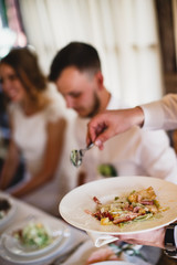 A male hands are holding a plate with a salad, and a spoon. The guests sitting behind a table are at the background.