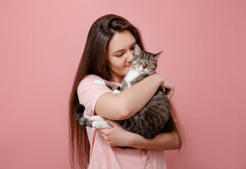 young attractive woman hugging cat in hands, pink background, girl kissing cat