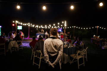 Waitress is standing at the foreground. A lot of tables with many guests sitting behind them are at...