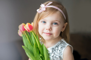 Obraz na płótnie Canvas Little blonde girl with a bouquet of tulips., pressed to the colors in a white dress with a ribbon in her hair, horizontal image, look at the camera, with a gentle smile