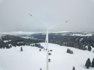 Aerial view of wind turbine in snow covered landscape in Swizterland. Tall pylon in fog, fir trees in the background.