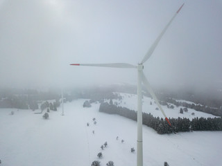 Aerial view of wind turbine in snow covered landscape in Swizterland. Tall pylon in fog, fir trees in the background.