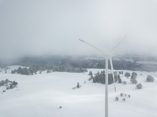 Aerial view of wind turbine in snow covered landscape in Swizterland. Tall pylon in fog, fir trees in the background.