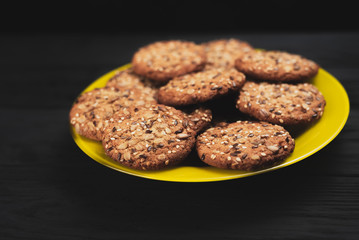 Oatmeal cookies with seeds and sesame seeds on a yellow plate, on a black wooden background