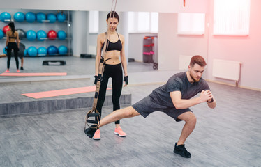 Fototapeta na wymiar Strong beardy trainer doing exercise with TRX and a woman in sportswear looking at him in gym. Pretty female standing near handsome man doing legs training on TRX. Sport concept