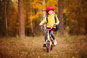 cheerful blonde babe 6 years old in a red hat yellow sweater in autumn riding a bike in the park or in the woods