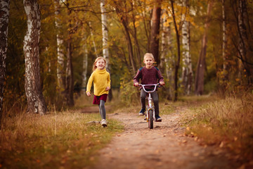 happy baby, baby boy 7 years riding a bicycle, and his little sister's sister, a little girl in a red hat runs with him along the park in the golden autumn