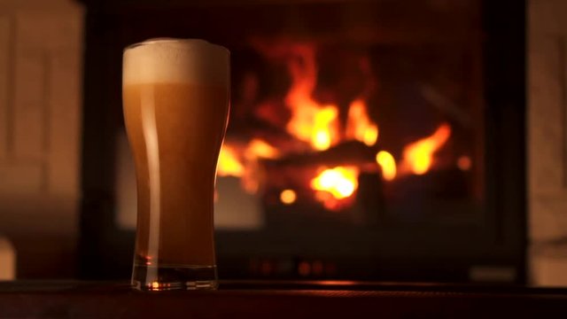 Full shot pouring stream into big glass bocal of refreshment beer with foam and air bubbles. Cosiness winter countryside atmosphere with yellow and orange flame of fireplace slow motion